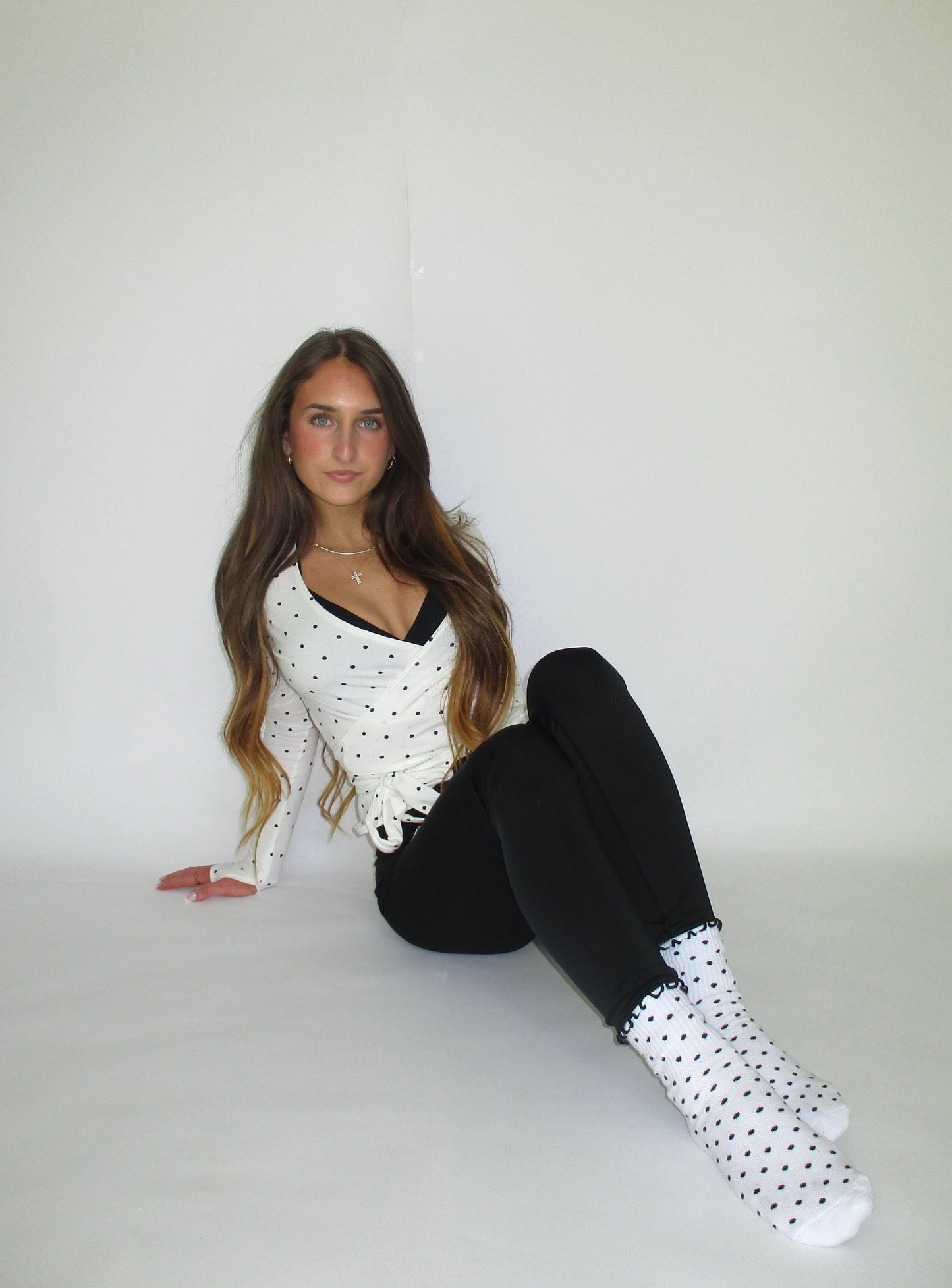 Woman sitting on a white surface wearing a polka dot outfit with black leggings and white shoes.